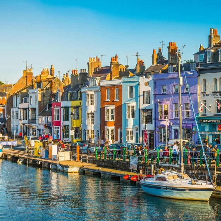 People walking up and down the water front of a busy seaside town lined with colourful terraced houses and boats anchored in the harbour