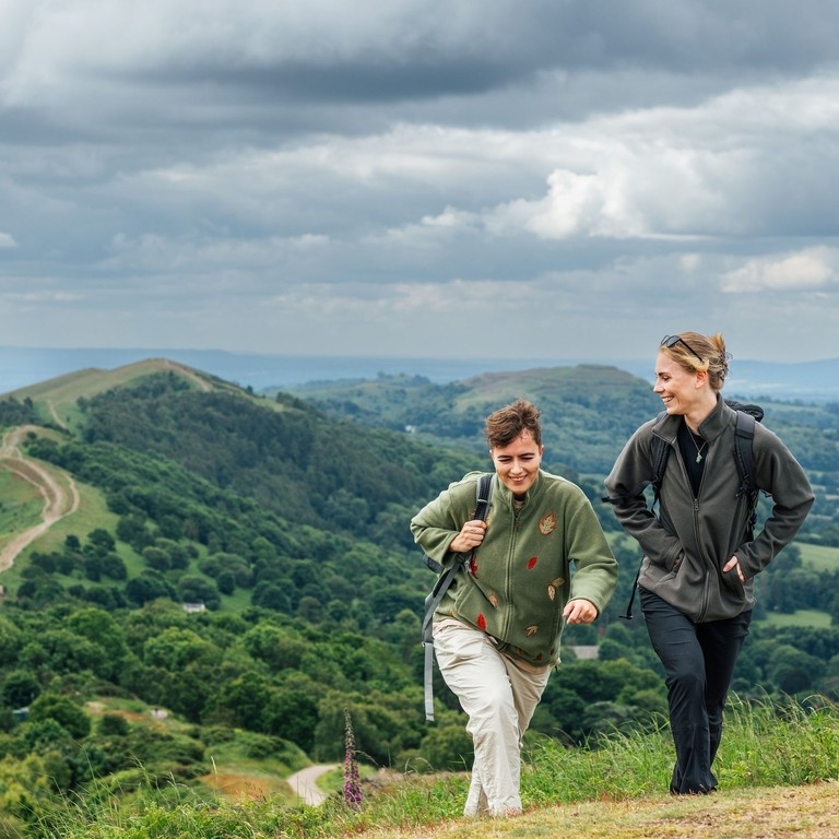 Two women hike carrying rucksacks along green hills on a cloudy day