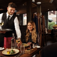 A smartly dressed staff member serving wine and food to a couple in a dining room 