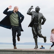 View of man, female child and statue of a man in front of the sea