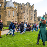 A group of people lined up for a Broomstick Training lesson with the wizarding professors by a large castle.
