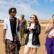 A group of friends walk on the beach with ice creams, a statue in the background
