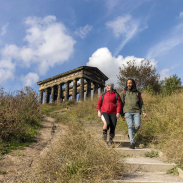 Two people wearing outdoor clothing, hiking past a stone monument, surrounded by greenery and blue skies.